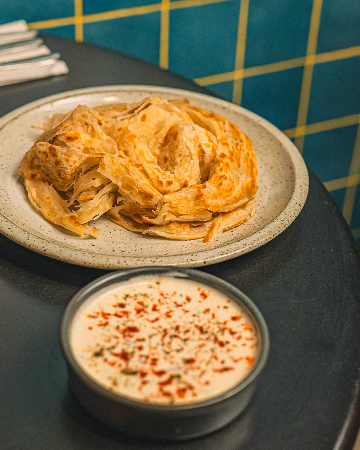 Flaky layered paratha on a plate, with a bowl of raita