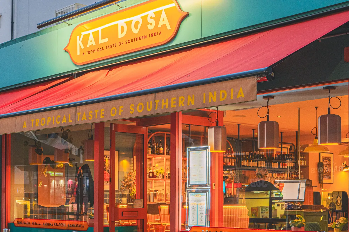 Kal Dosa's shopfront at dusk: the orange sign on the teal fascia, a red awning, and the room glowing through red-framed windows