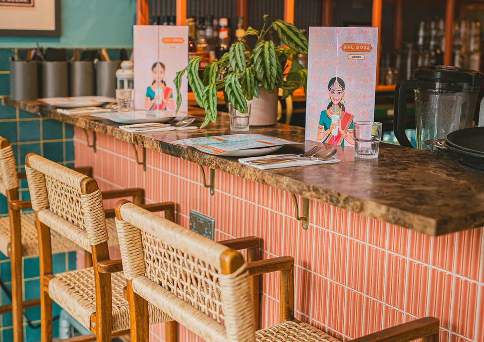 Rattan bar stools at a pink-tiled bar, under shelves of bottles